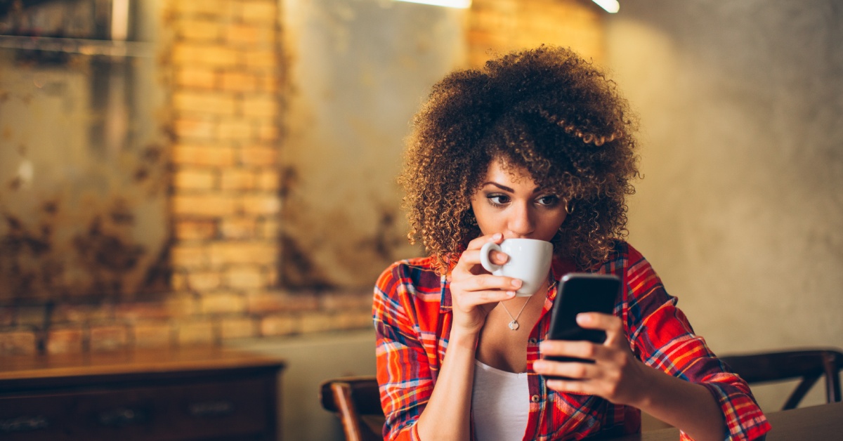 A person in a plaid shirt drinking a cup of coffee at home and looking at the cell phone in their hands.