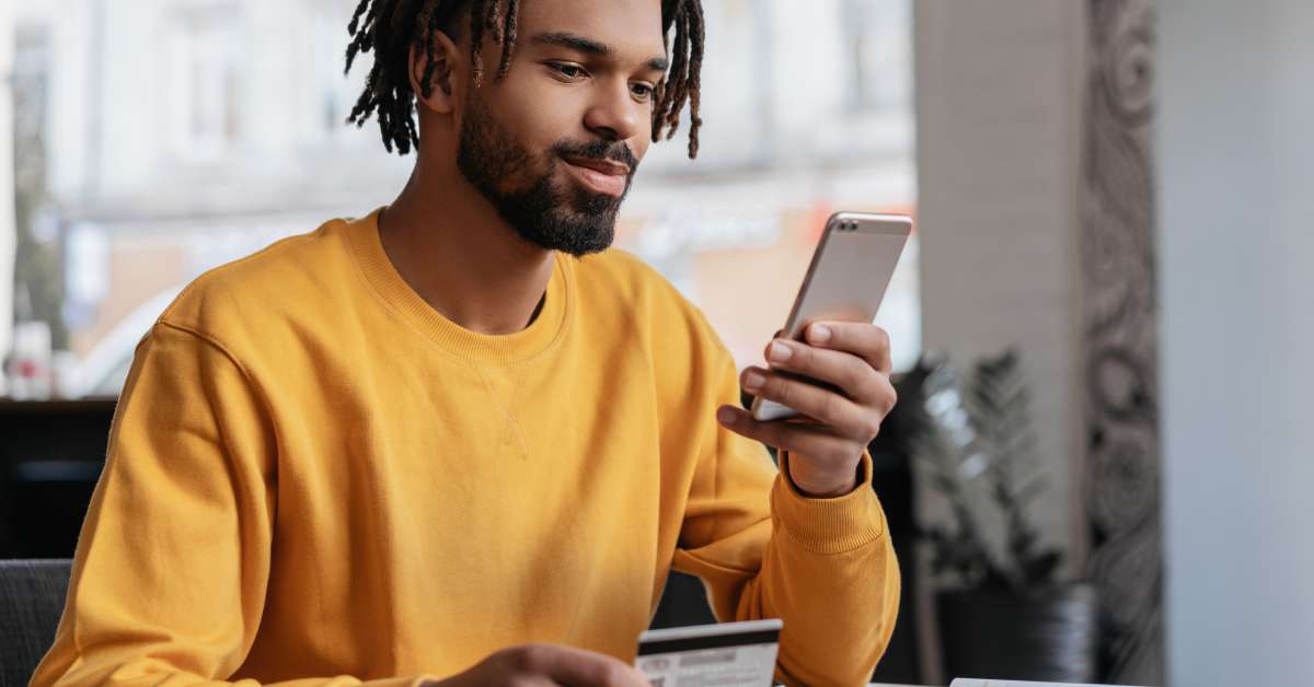 A person wearing an orange sweatshirt is holding their cell phone and browsing it in the middle of a large room.