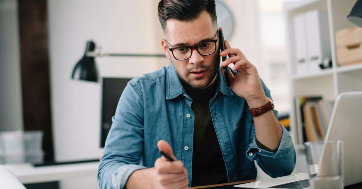 A person wearing glasses at a workstation anxiously listens to a phone call while holding a pen in one hand.