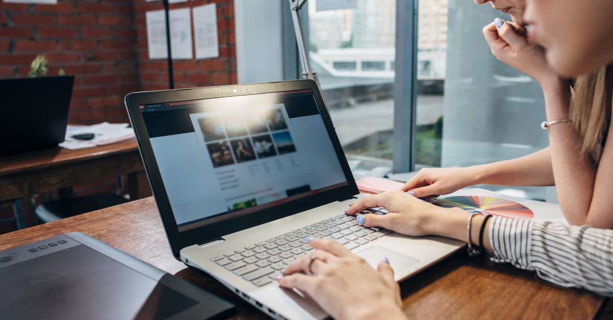 A person is using a grey laptop in a home office while another person is next to them and reading a pink note on a desk.