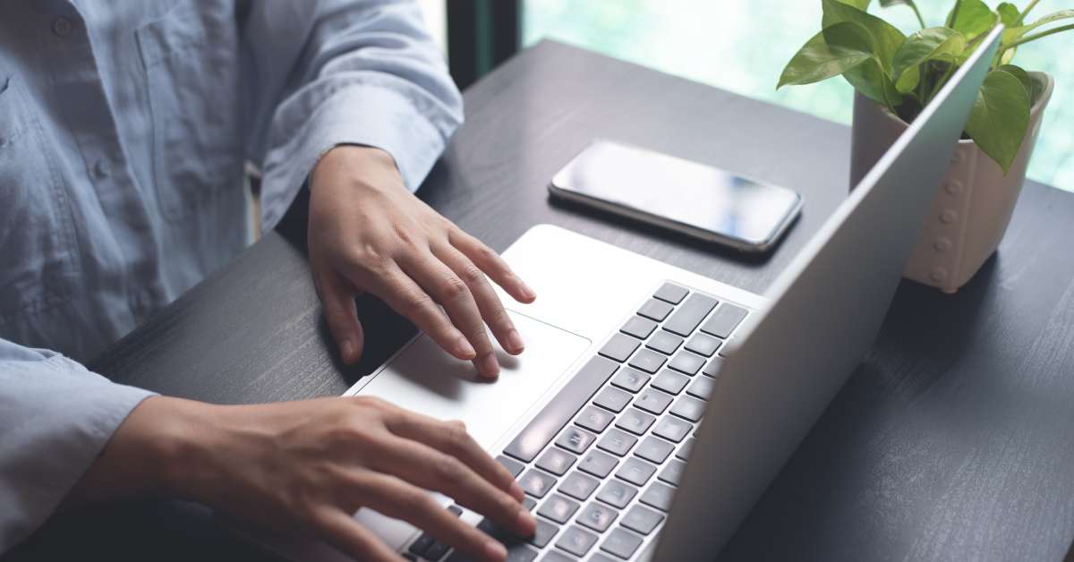 A person sitting at a dark brown desk is typing on a laptop that is sitting next to a potted plant and a cell phone.