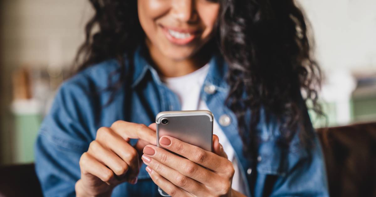 A person with their sleeves rolled up and smiling while scrolling through an app on their small silver cell phone.