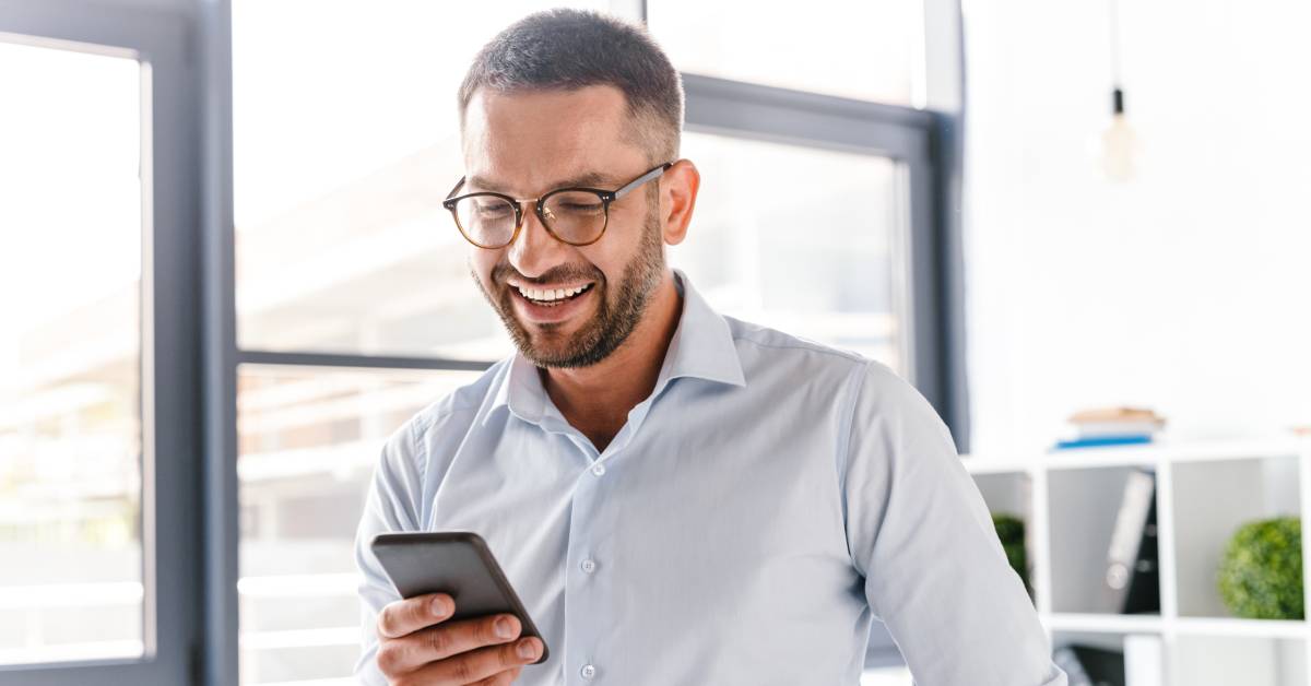 A person wearing glasses and casual yet professional clothing. They smile while looking down at their cell phone.