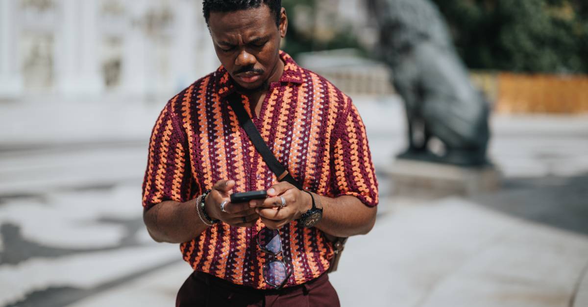 A person with a concerned look on their face is looking down at the cell phone they’re holding while standing outside.