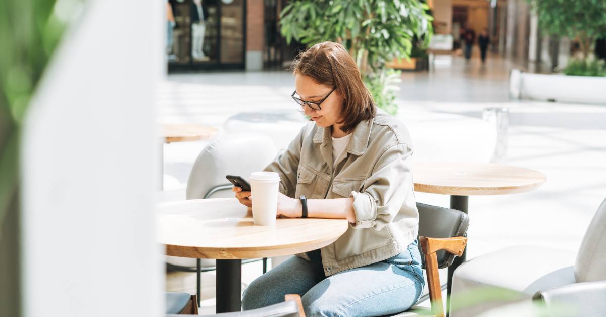 A person is sitting down at a round wooden table in a large public space while using their cell phone.