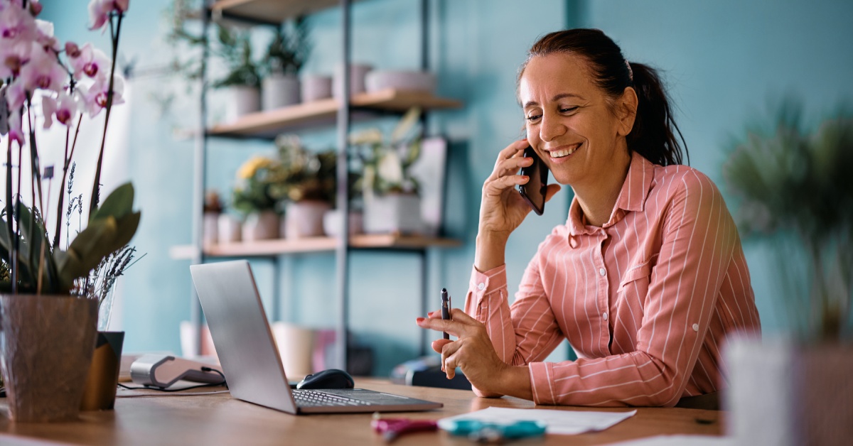 A woman is wearing a pink shirt with white stripes and is smiling while talking on their cell phone.