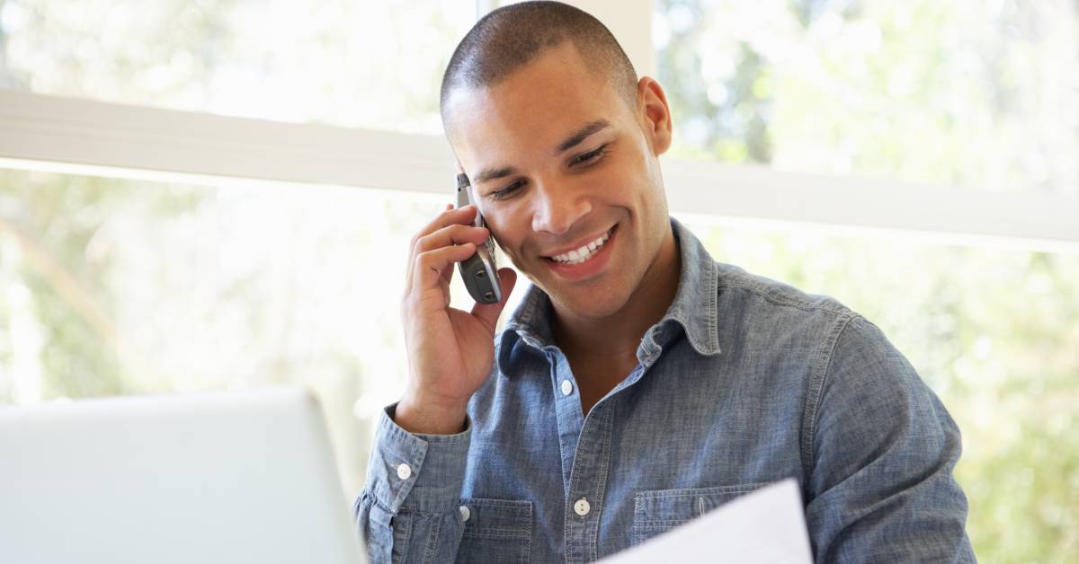 A man is smiling and talking on their cell phone while observing a document that is sitting next to their laptop.