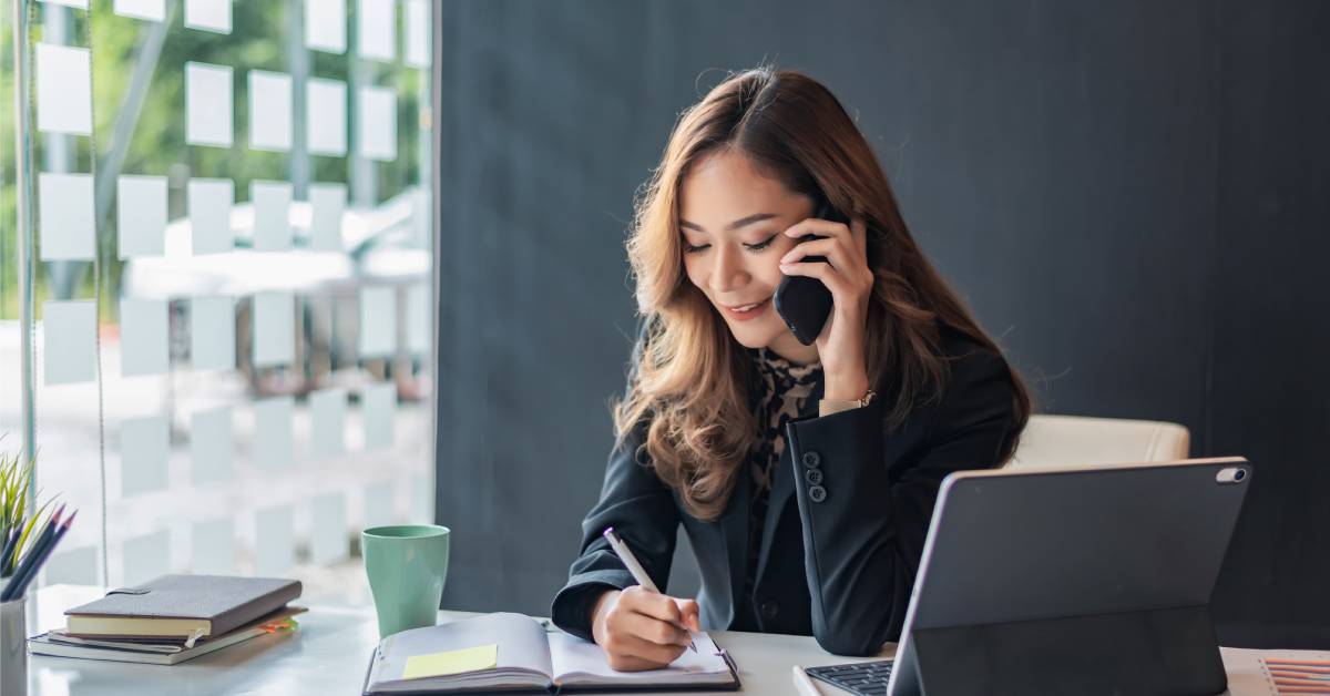 A person is talking on the phone while writing in a notebook next to their computer and a green cup.