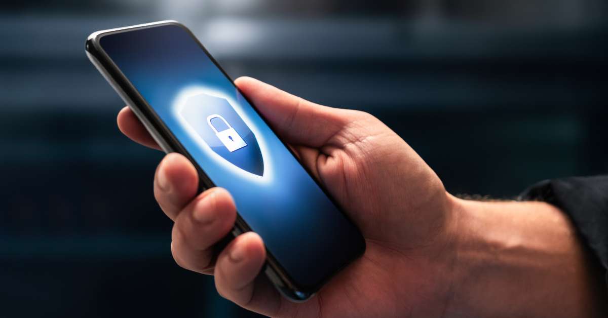 A person is holding a cell phone that is displaying a blue shield symbol with a lock at its center on the screen.