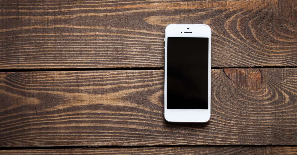 A small white cell phone with a dark blank screen is sitting alone on a large dark brown wooden table.