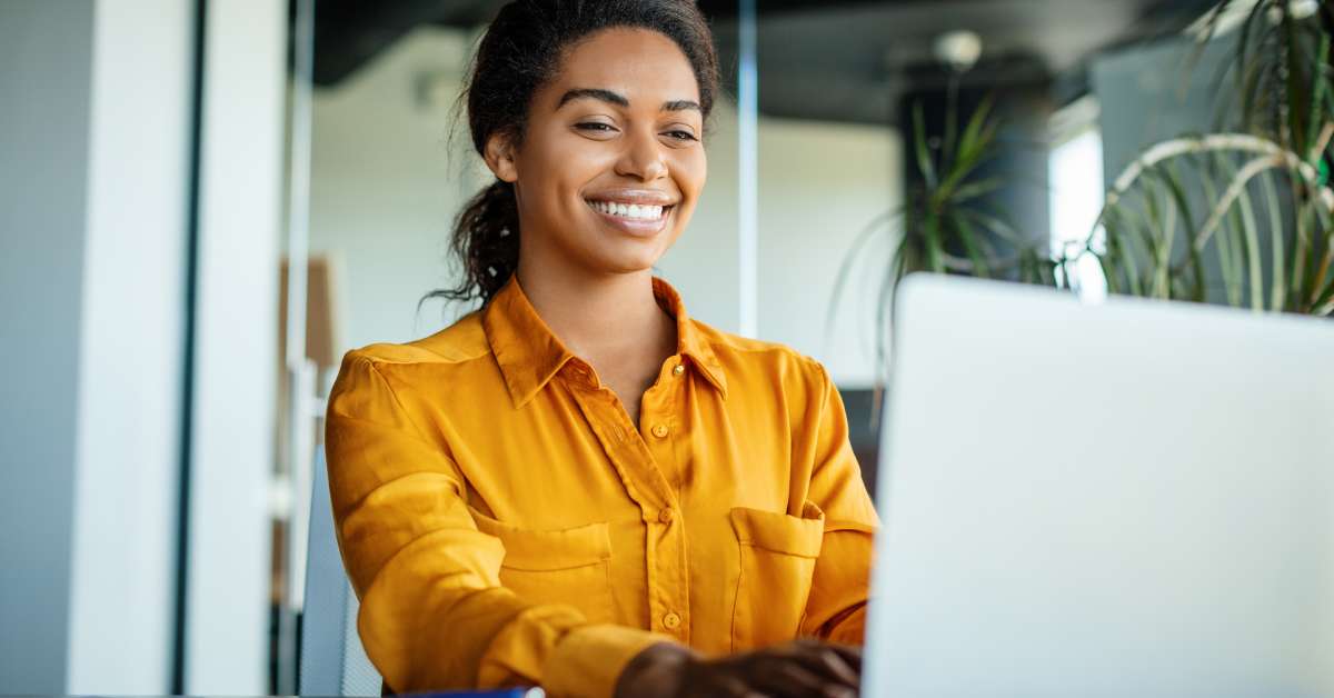 An employee wearing a bright orange shirt is sitting in an office and smiling as they type on a computer.