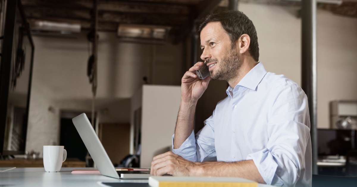 An employee smiles while talking on their cell phone in an office. They are sitting at a large desk with a laptop.