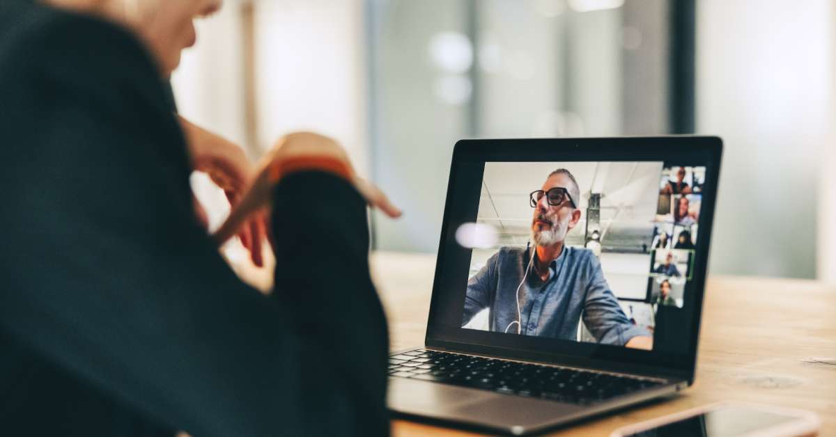 A person in a black shirt is sitting at a wooden table and hosting a video conference on their laptop.