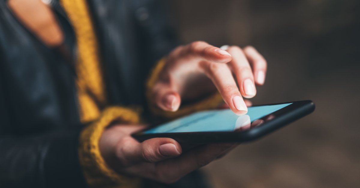 A person wearing a black jacket over a yellow long-sleeve is scrolling through their cell phone outside.