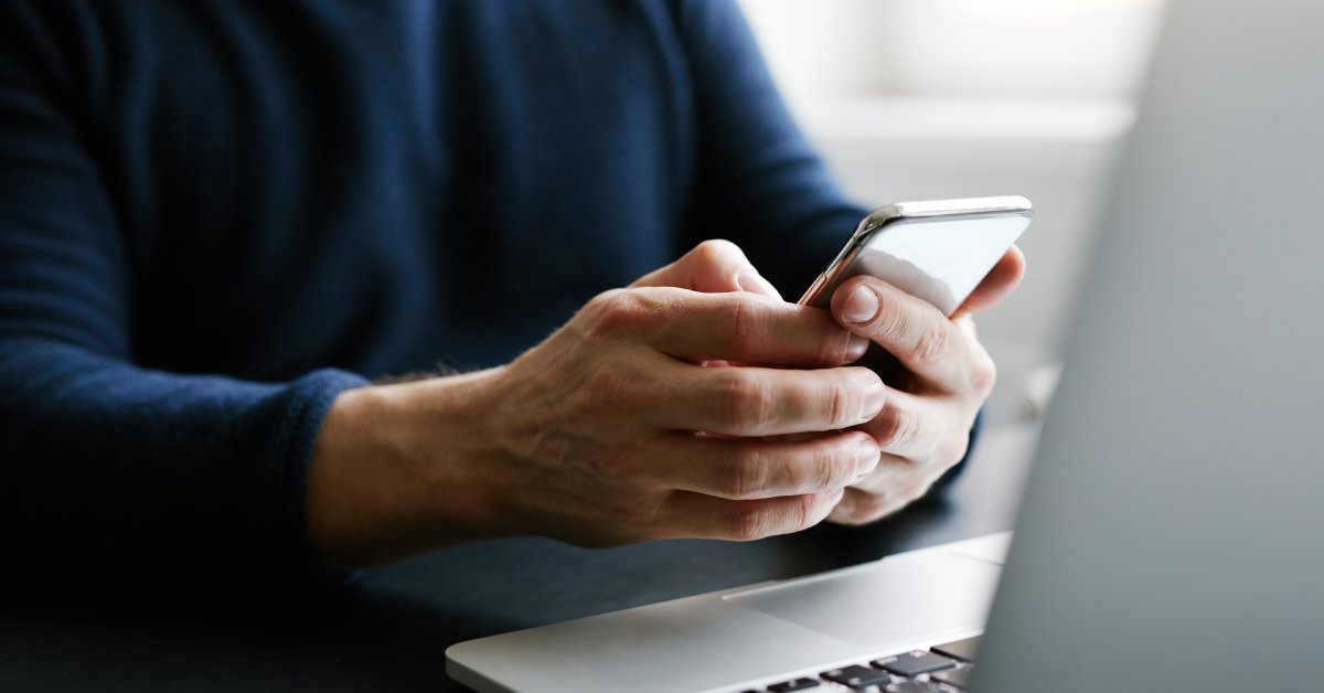 An individual is sitting and holding their cell phone over their laptop that is open in front of them.