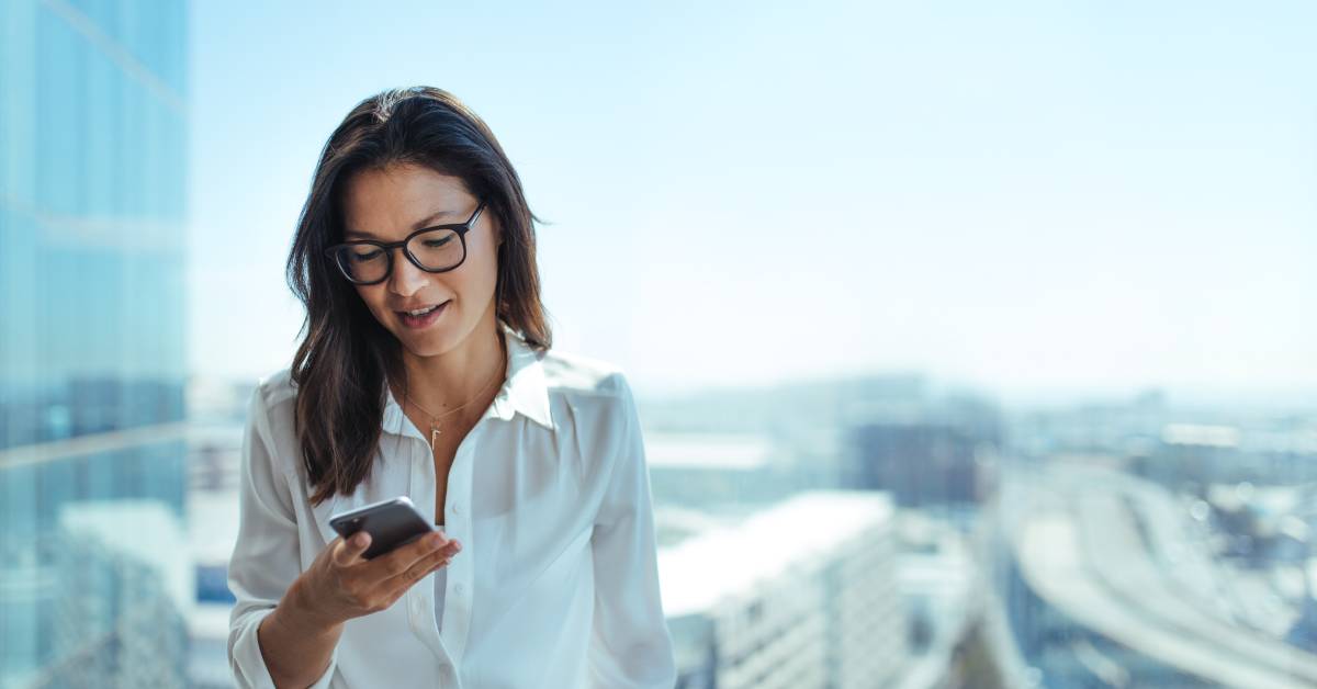 An employee wearing glasses and a white shirt is standing outside during daytime while looking down at her cell phone.