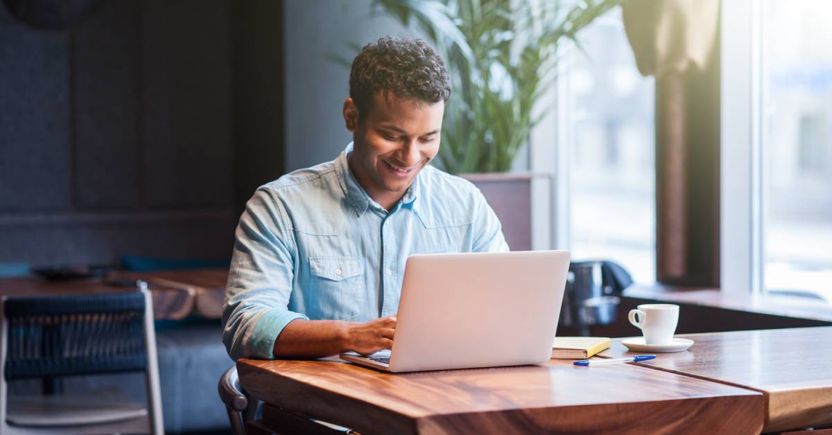 A man is smiling and working on his laptop while seated at a modern wooden table.