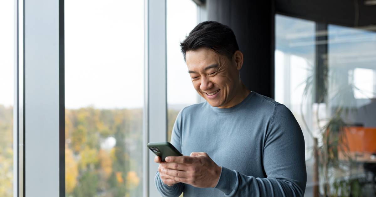 A man standing next to large windows in an office is smiling while texting on his cell phone.