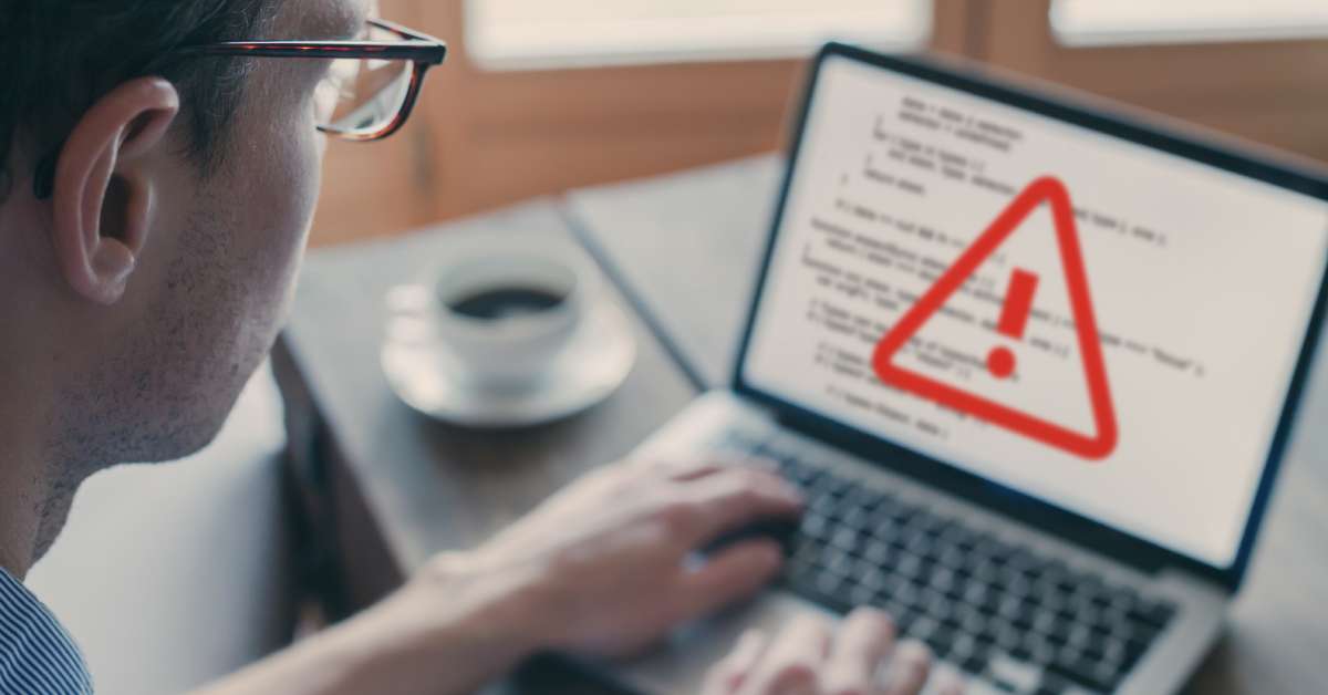 A person is looking at a computer screen that has a red warning symbol on it. The computer is next to a cup of coffee.