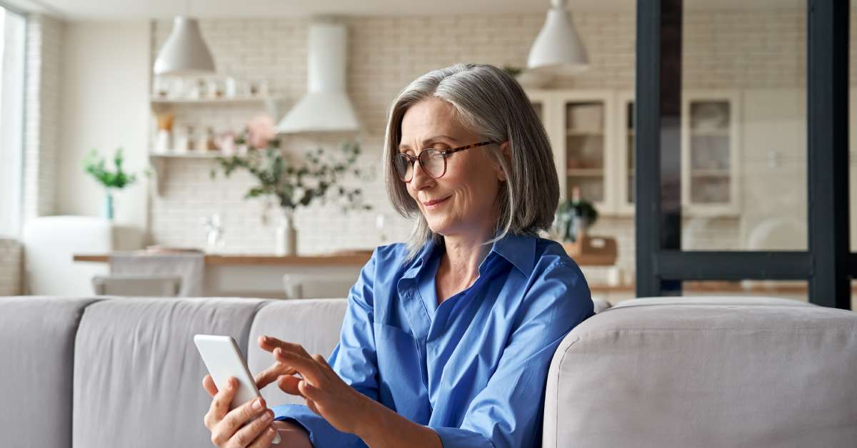 A homeowner wearing a blue shirt and glasses sitting in their home and using their cell phone to browse the internet.