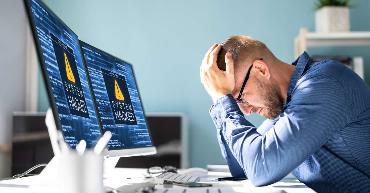 Two computer monitors in an office read “System Hacked.” An employee sits in front of the screens, frustrated.