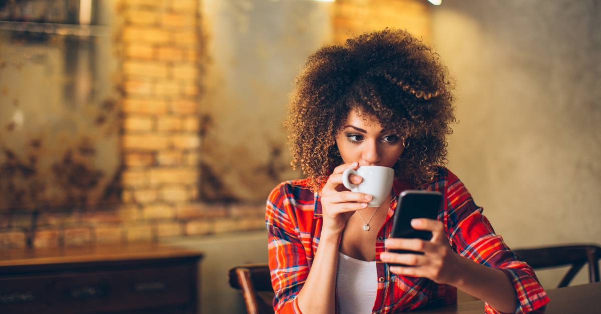 A homeowner sits alone in their brightly lit kitchen. They are sipping a cup of coffee while browsing their cell phone.