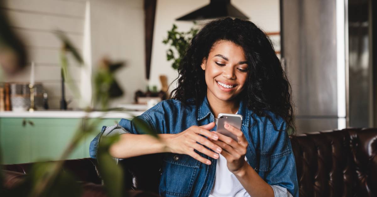 A person in a blue jacket and white shirt is smiling while sitting on a couch and looking at their cell phone.