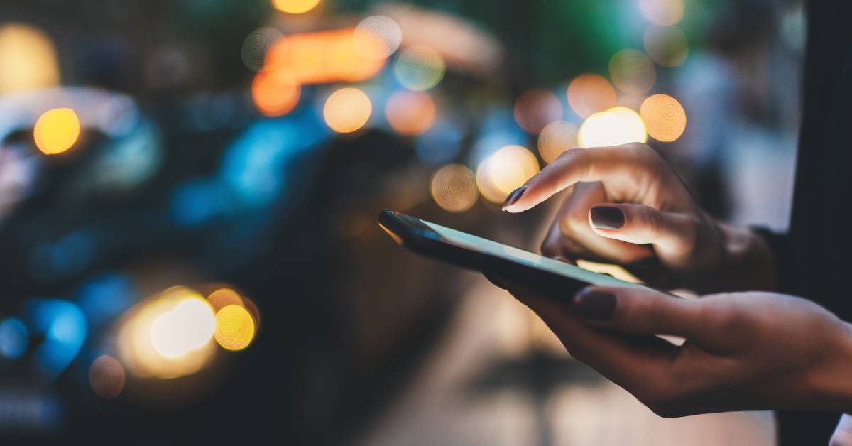 A cell phone user with dark nail polish is standing on the street at night while browsing a mobile device.