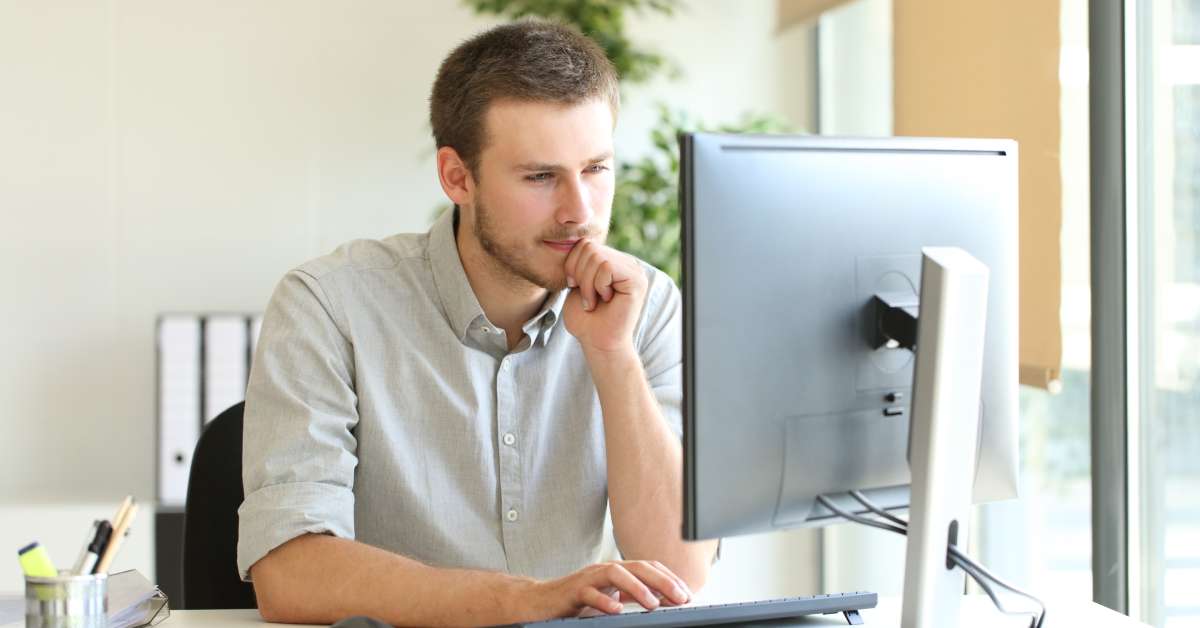 A worker sits at their desk and narrows their eyes while looking at a computer. The desk is next to a large window.