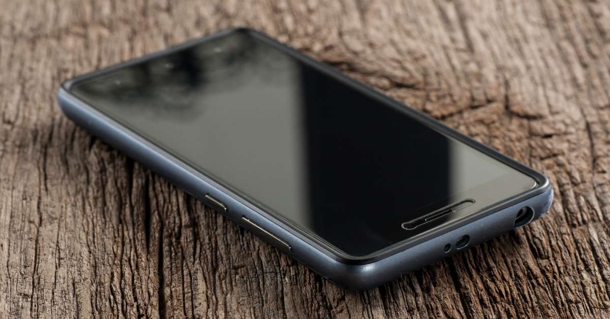 A black cell phone is sitting on a brown wooden table by itself. The phone's screen is not turned on.