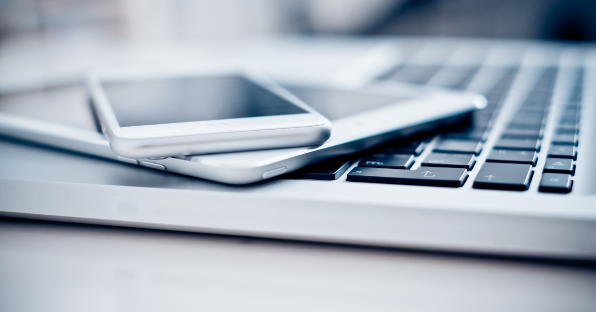 A tablet is lying across the keyboard of a laptop, and a cell phone is on top of the tablet. All three devices are gray.