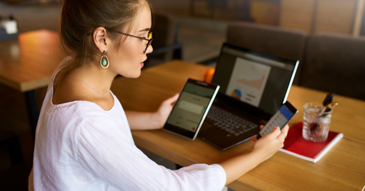 A person is sitting at a desk while holding a cell phone in one hand and a tablet in the other. A laptop is on the desk.