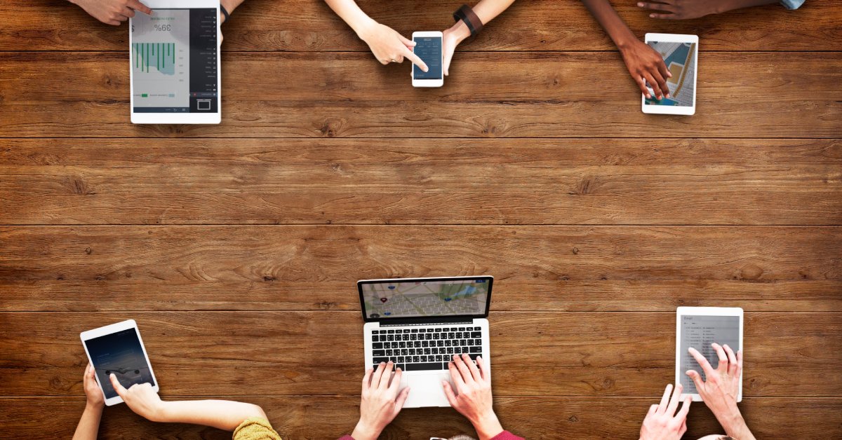 Six people are at a wooden table, and four of them are using tablets. The other two have a cell phone and a laptop.