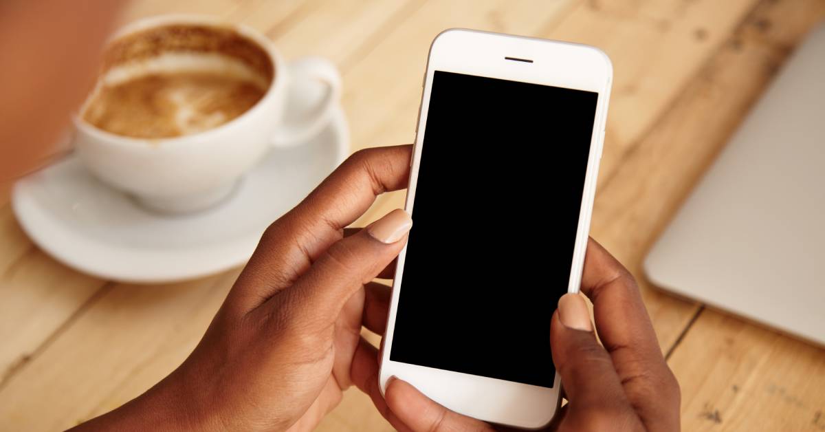 A person is holding their cell phone over a wooden table. A coffee cup and a closed laptop are on the table.