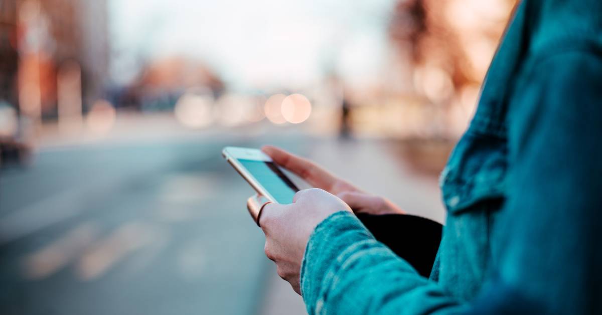  A cell phone user is browsing their device while standing next to the street in the middle of the day.