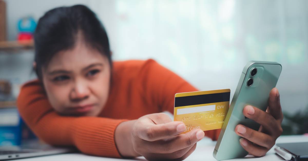 A person wearing an orange long-sleeved shirt leans over and holds a debit card next to their cell phone.