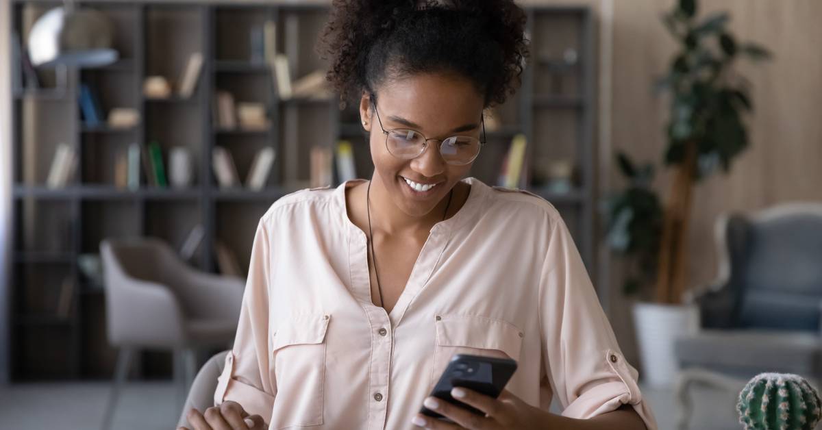 A person wearing glasses and a pink shirt smiles while sitting at their desk and looking at their cell phone.