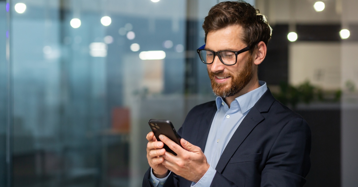 A person wearing a suit and thick-rimmed glasses stands inside while staring down at their cell phone.