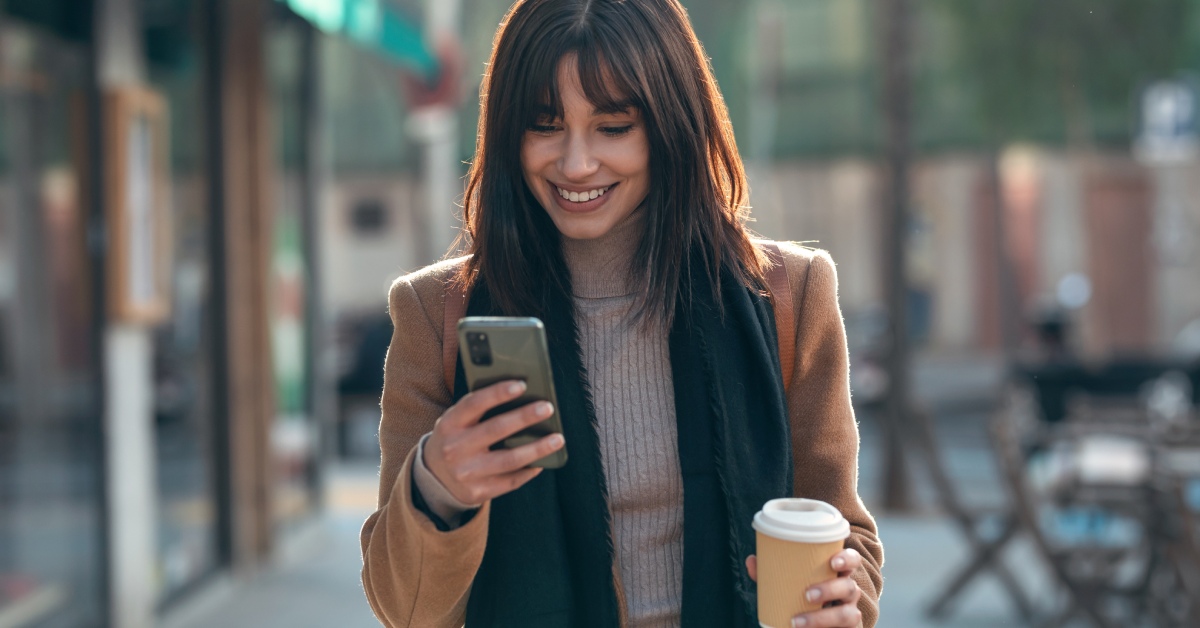 A person smiles outside while looking at the phone in their right hand. They are carrying coffee in their left hand.