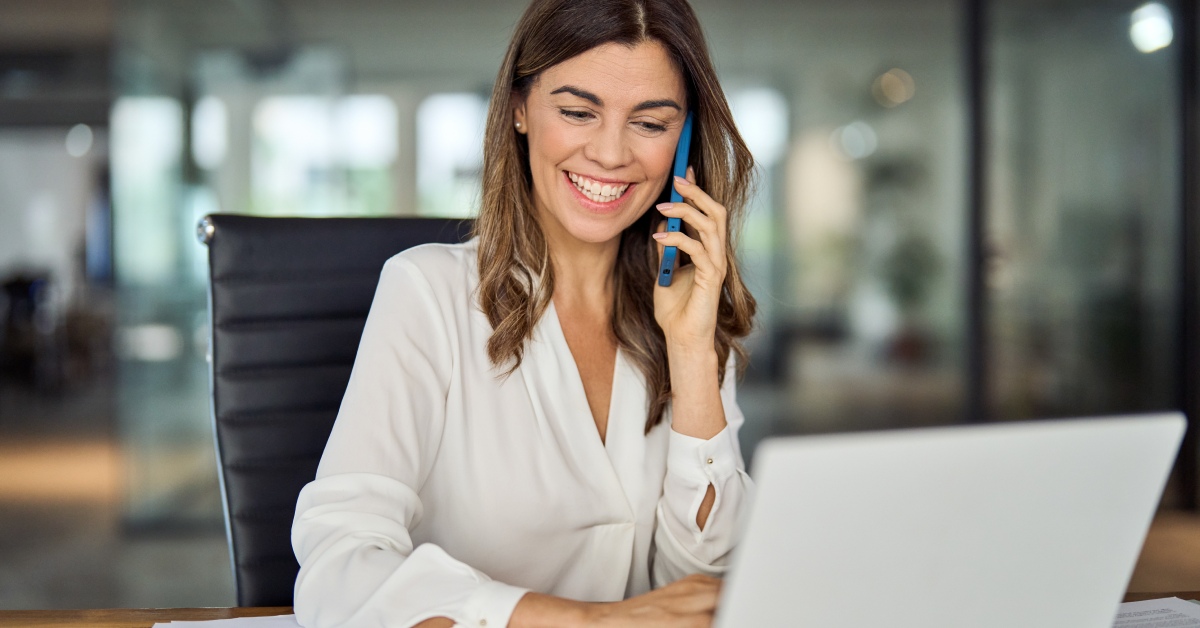 A professional is sitting in the office and smiling while having a phone call on their blue cell phone.