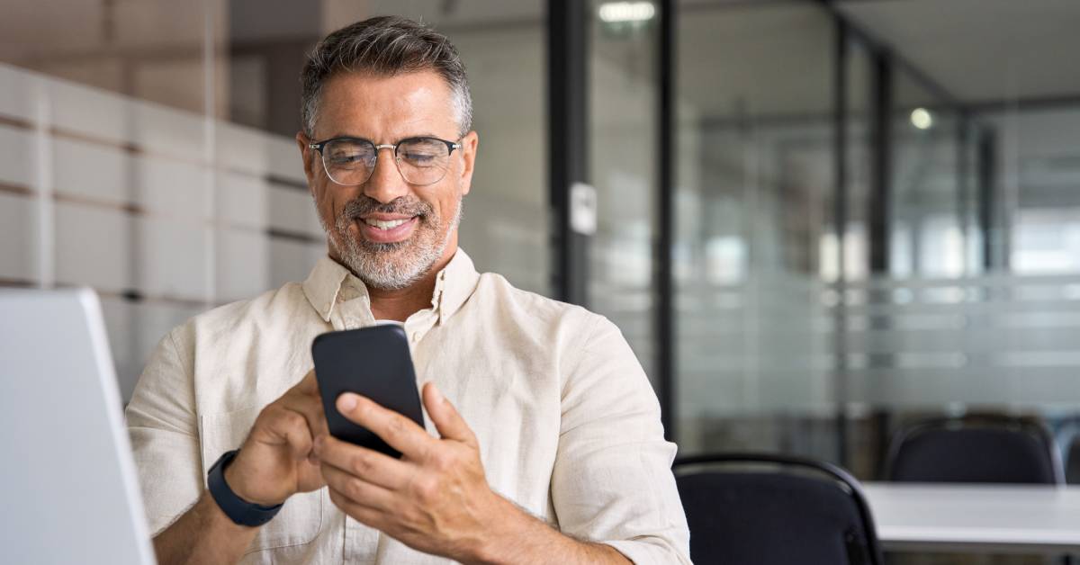 A person wearing glasses and a white shirt is smiling while sitting and scrolling through their cell phone.