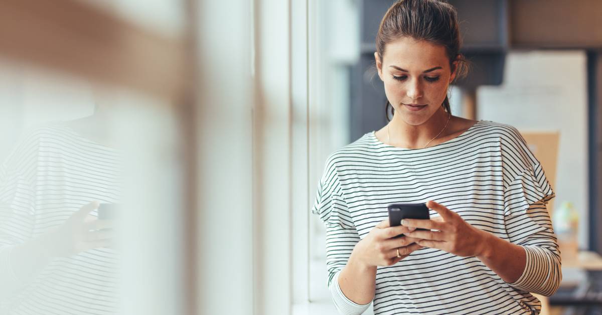 A person wearing a striped shirt is looking down at the cell phone in their hands while standing next to a large window.