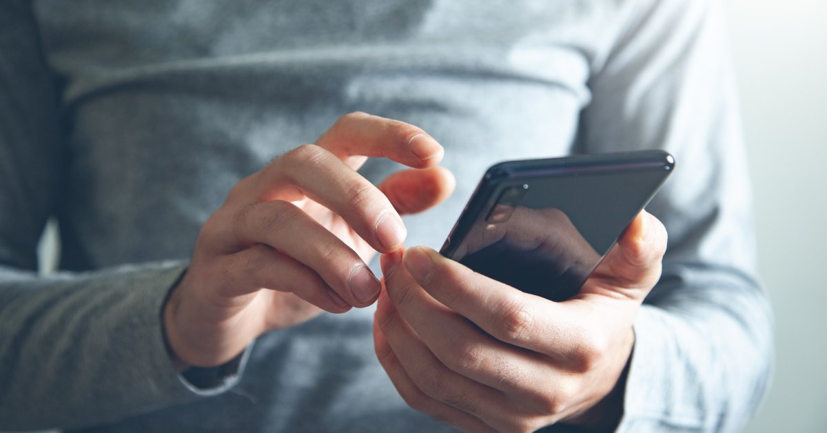 A person wearing a gray long-sleeved shirt is using their finger to tap the screen of their black cell phone.