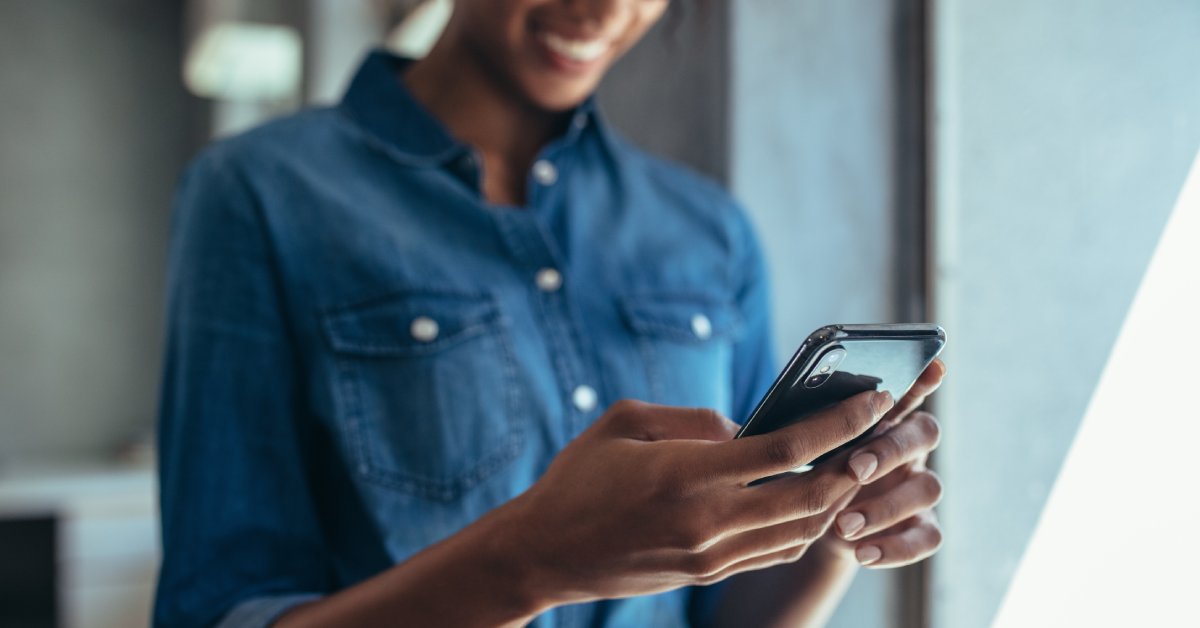 A smiling person wearing glasses and a blue jean jacket who is looking down at the cell phone in their hands.