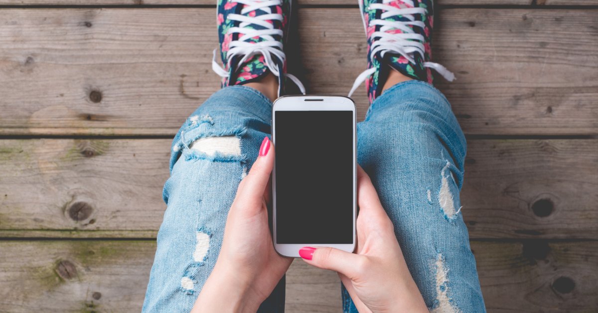 A person with pink nail polish is holding their cell phone over their torn blue jeans and colorful untied shoes.