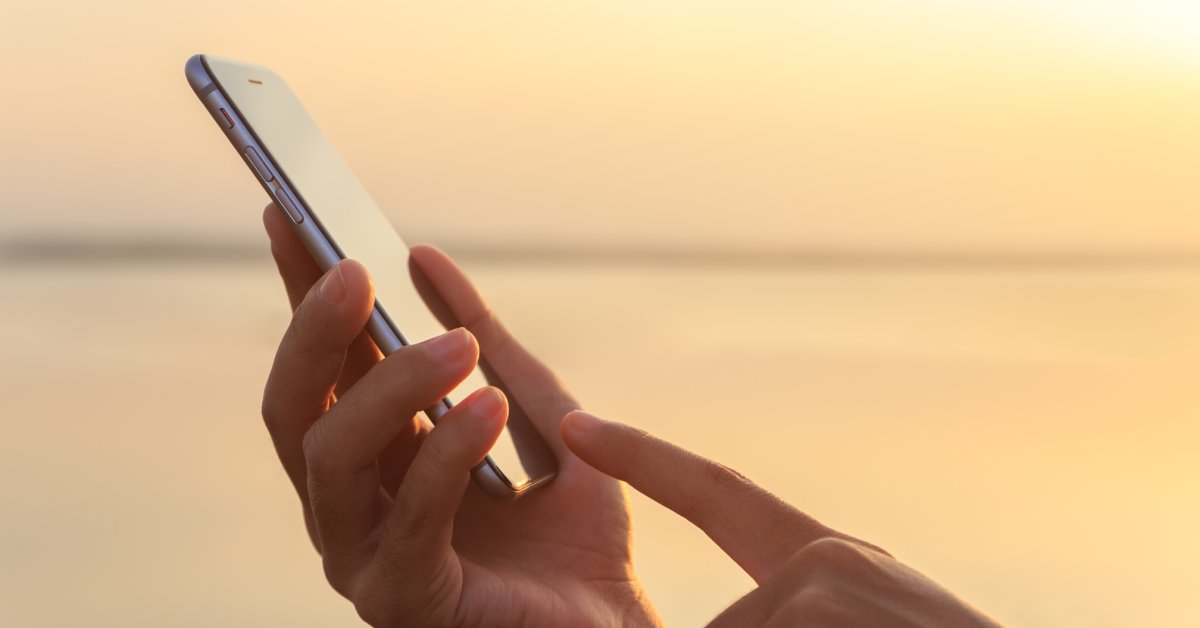 A person using their finger to tap on a cell phone screen. The sun is setting behind them and creating an orange glow.