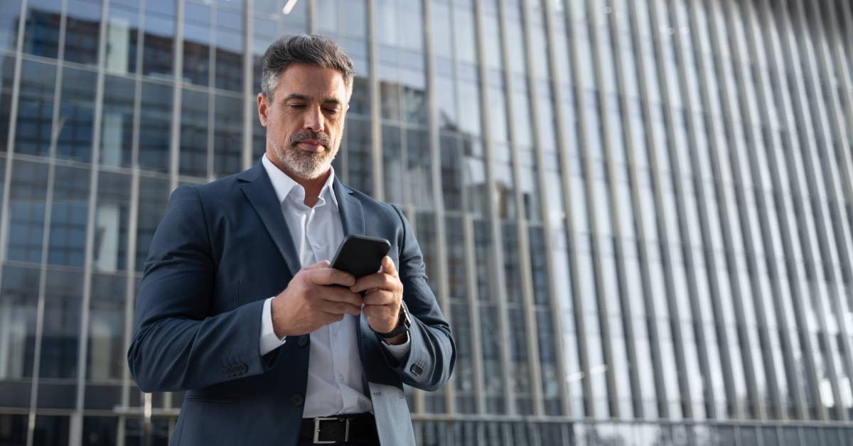 A person with a beard is standing alone outside of a large building and looking down at their cell phone.