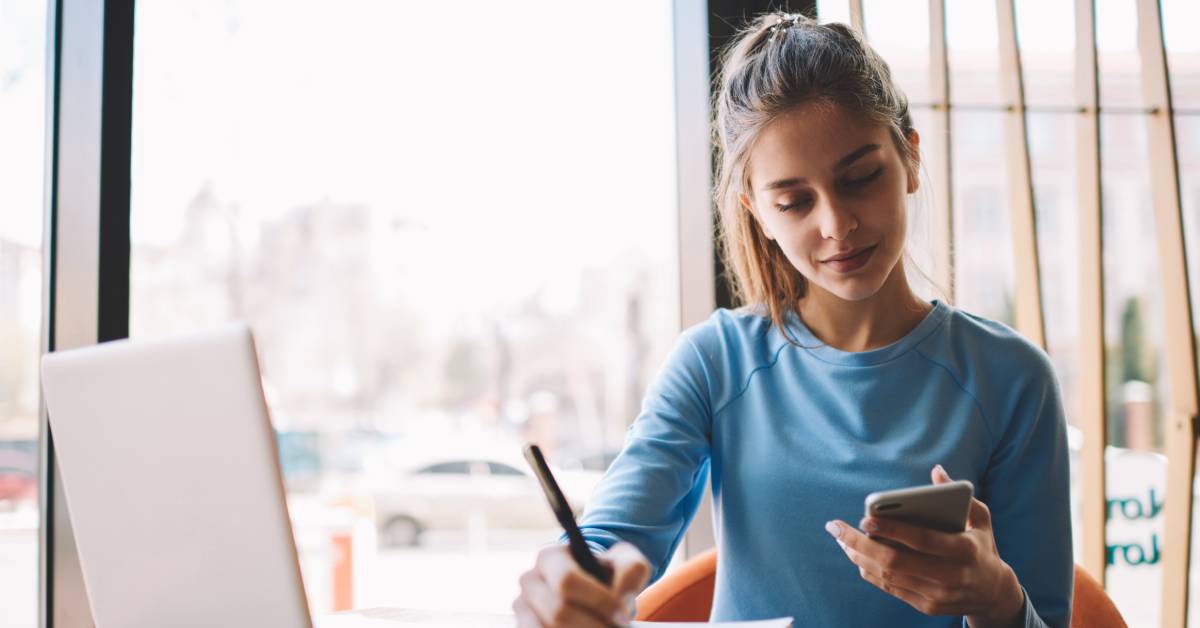 A journalist wearing a blue long-sleeved shirt is writing down notes while looking at the cell phone in their left hand.