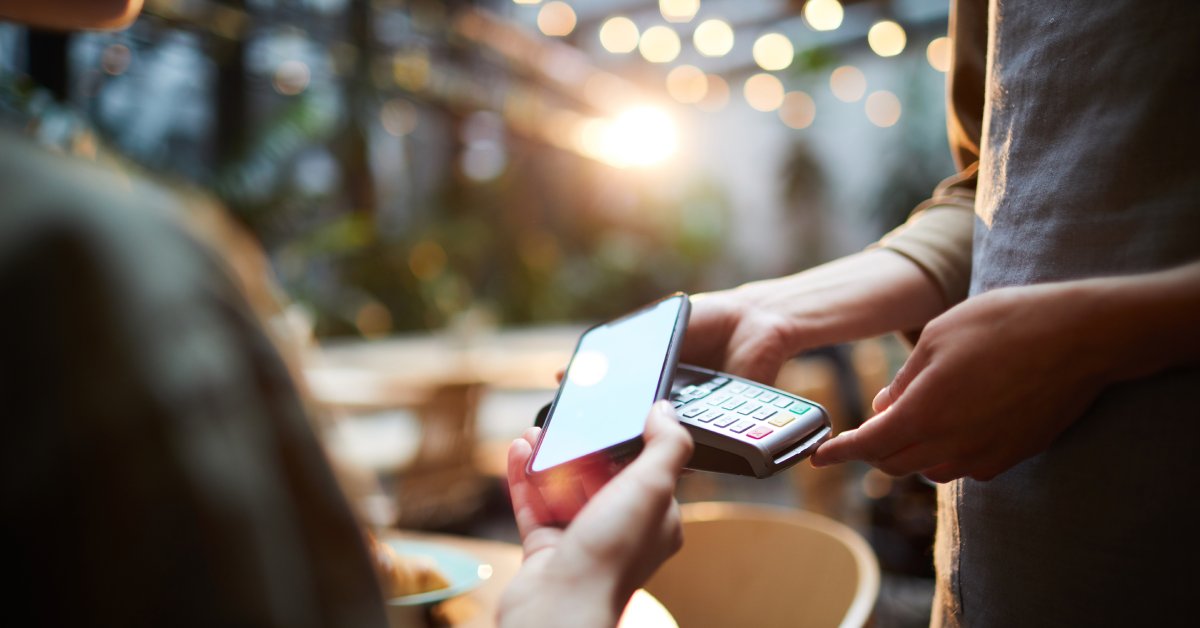 Someone sitting at a wooden table is holding their cell phone out toward someone who is holding a credit card reader.