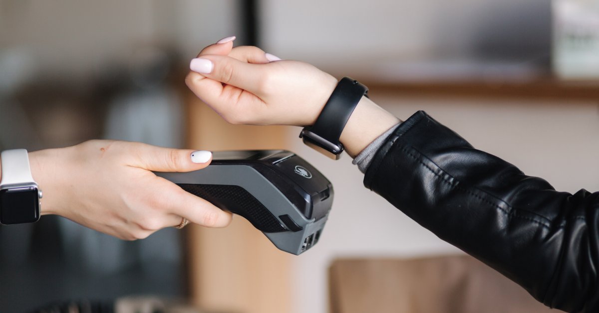 A person wearing a black jacket is holding the watch on their wrist above a black device someone else is holding.